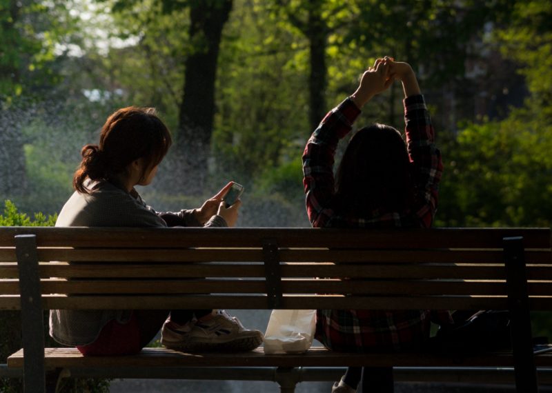 two girls sitting on a bench, one watching the phone and the other girl stretching